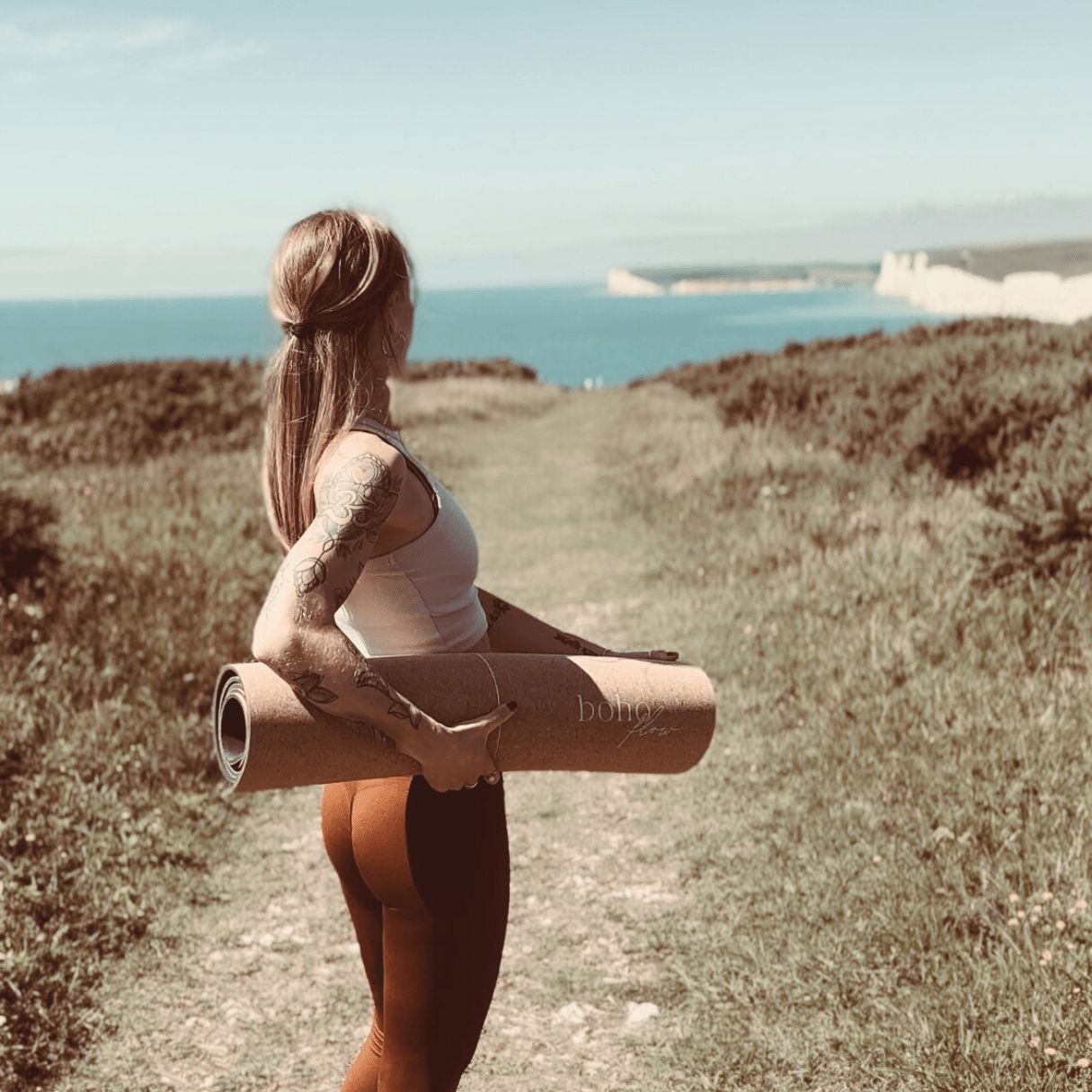 A girl carrying a cork yoga mat, standing on a cliff top with the ocean in the background.