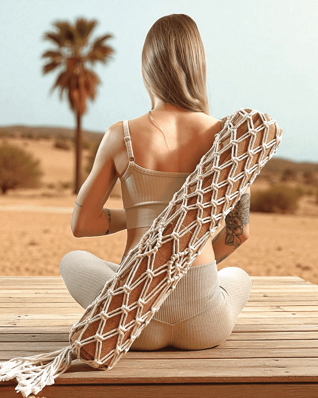 Woman sitting on a wooden bench in a desert setting with a macrame yoga mat bag around her shoulders.