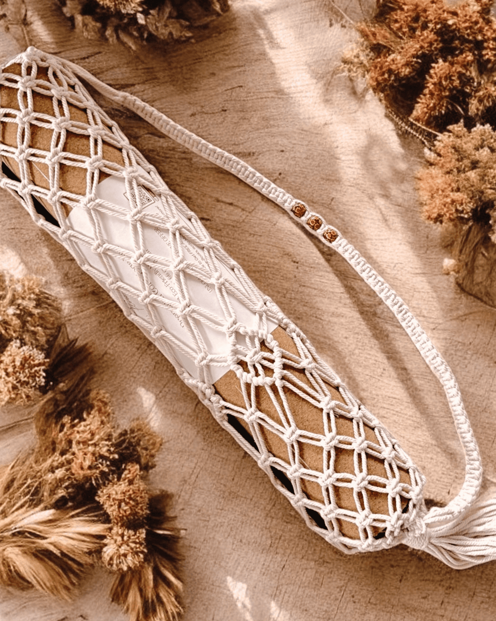 Macrame yoga mat bag containing a cork yoga mat lying on a wooden surface with dried plants