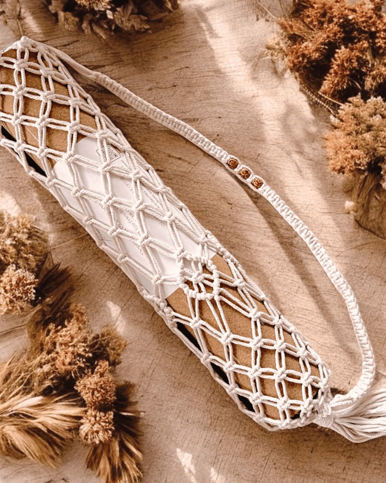 Macrame yoga mat bag containing a cork yoga mat lying on a wooden surface with dried plants