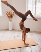 Person performing a handstand on a cork  yoga mat in a bright room with large windows and decorative plants.