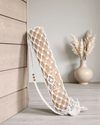 Macrame yoga mat bag leaning against a wooden wall with a vase of dried plants on a gray surface.