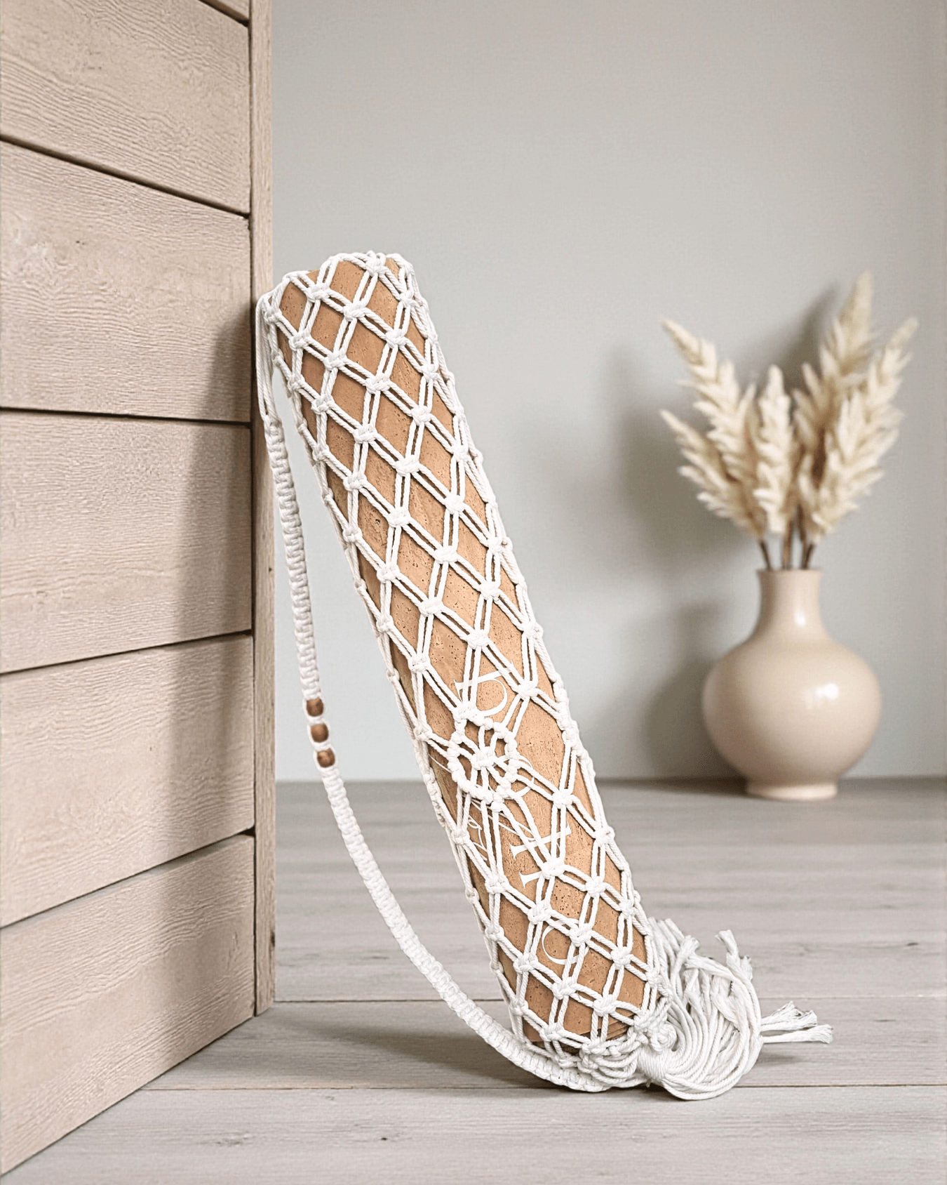Macrame yoga mat bag leaning against a wooden wall with a vase of dried plants on a gray surface.