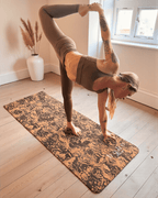 Person performing a yoga pose on a coffee and cork yoga mat in a room with a window and vase.