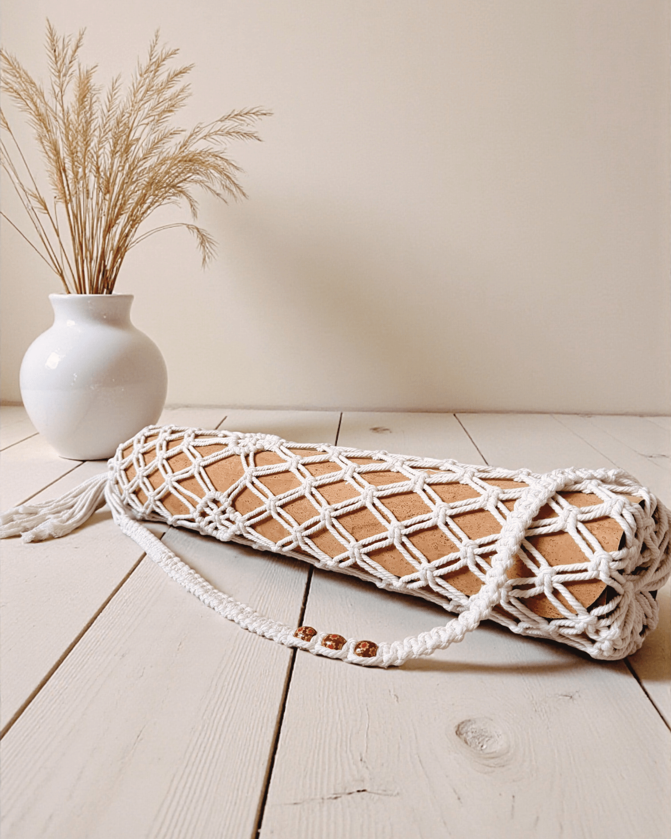 Macrame yoga mat bag on a wooden floor with a white vase containing dried plants in the background.