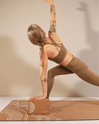 Woman in a yoga pose leaning on a cork yoga block on a yoga mat in a beige coloured room.