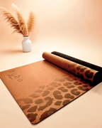 The Boho Flow Print Edition Cork yoga mat with leopard print design on a beige surface, rolled out and partially unrolled.