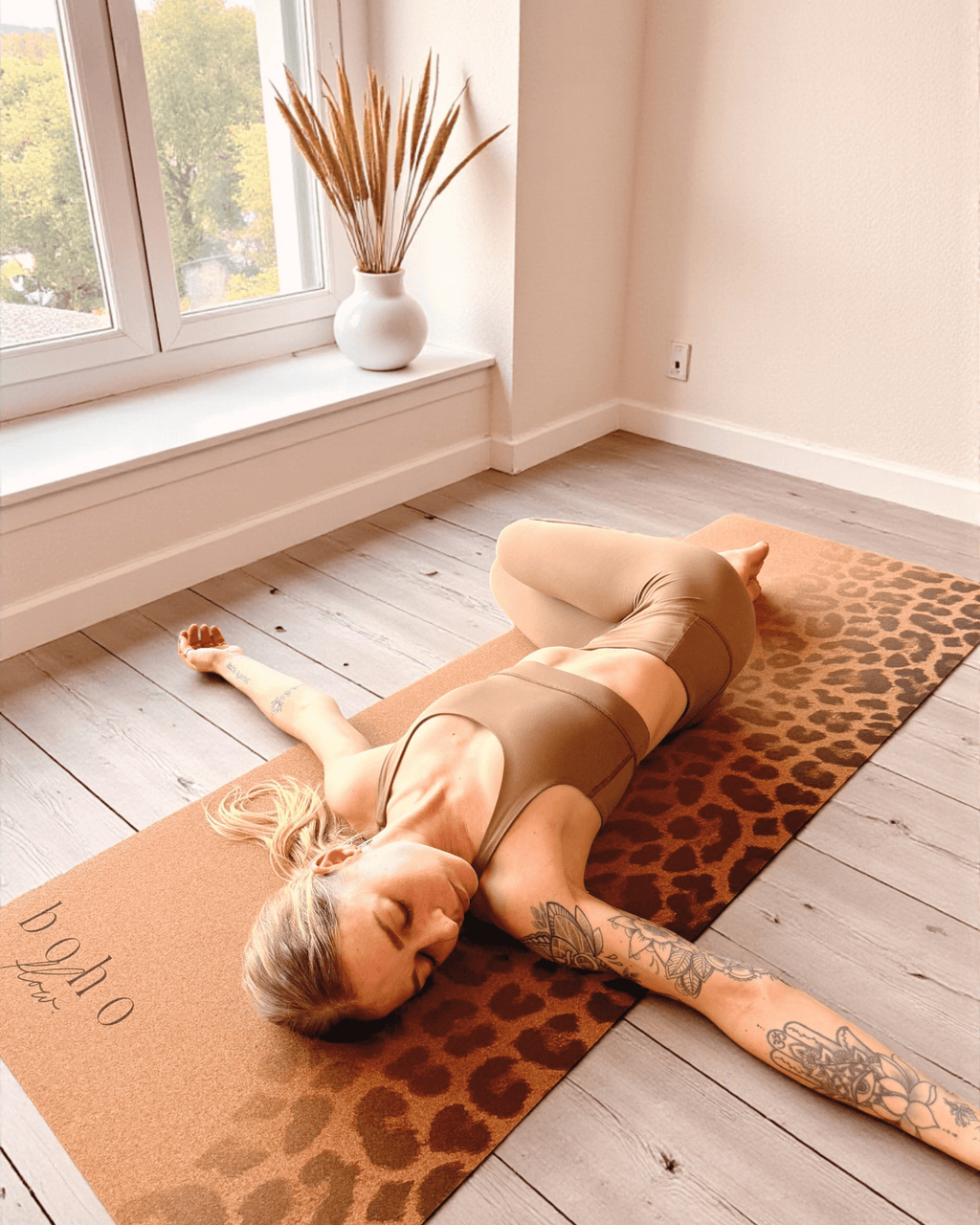 Person lying on a leopard print cork yoga mat in a bright room with large windows.