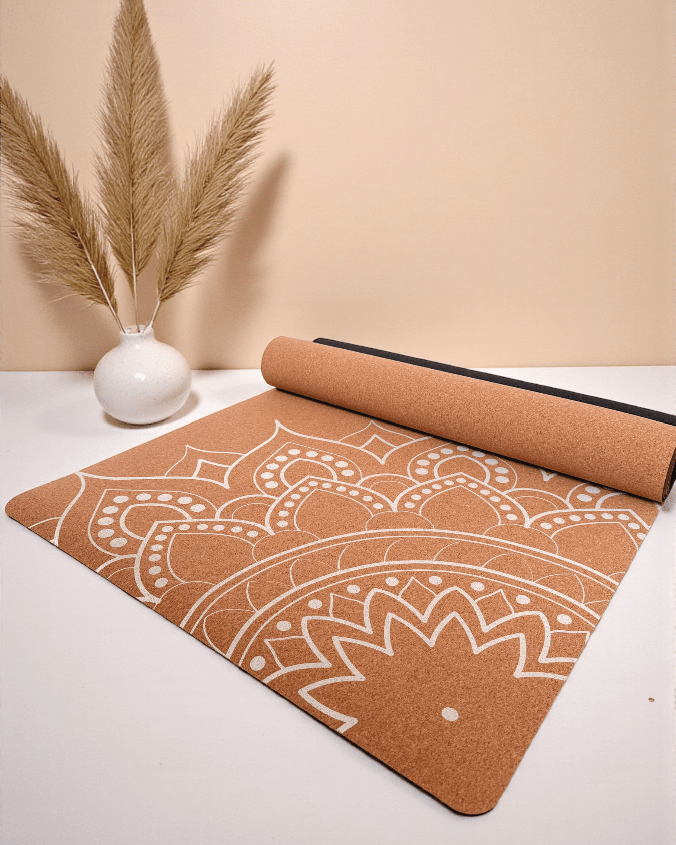 Cork yoga mat with white mandala pattern on a beige surface, accompanied by a small vase with dried plants.