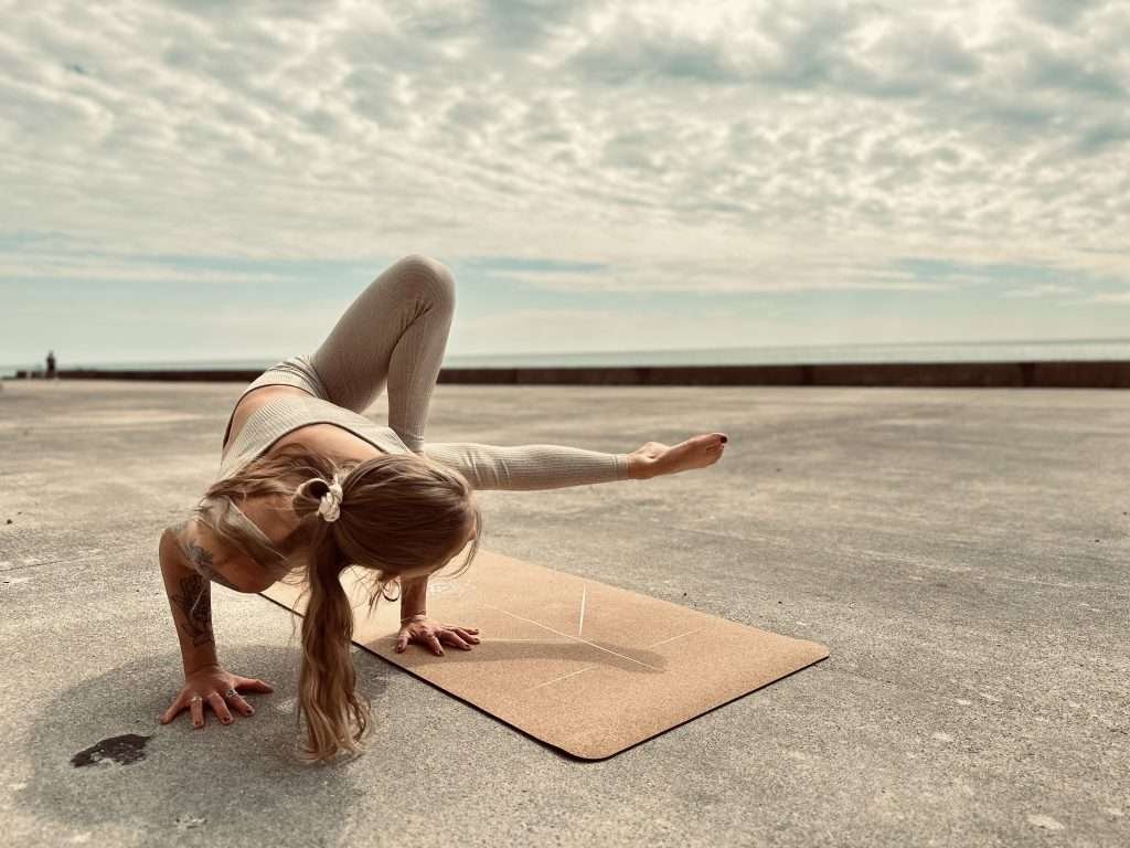 Girl practicing Grasshopper, the yoga arm balance, on a cork yoga mat outdoors by the sea.