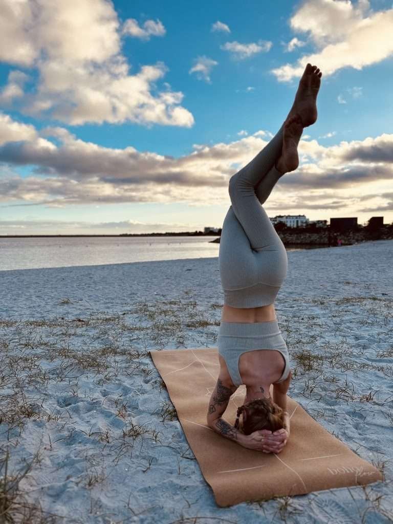 A girl practicing Garudasana leg Headstand variation outdoors on the beach on a cork yoga mat.